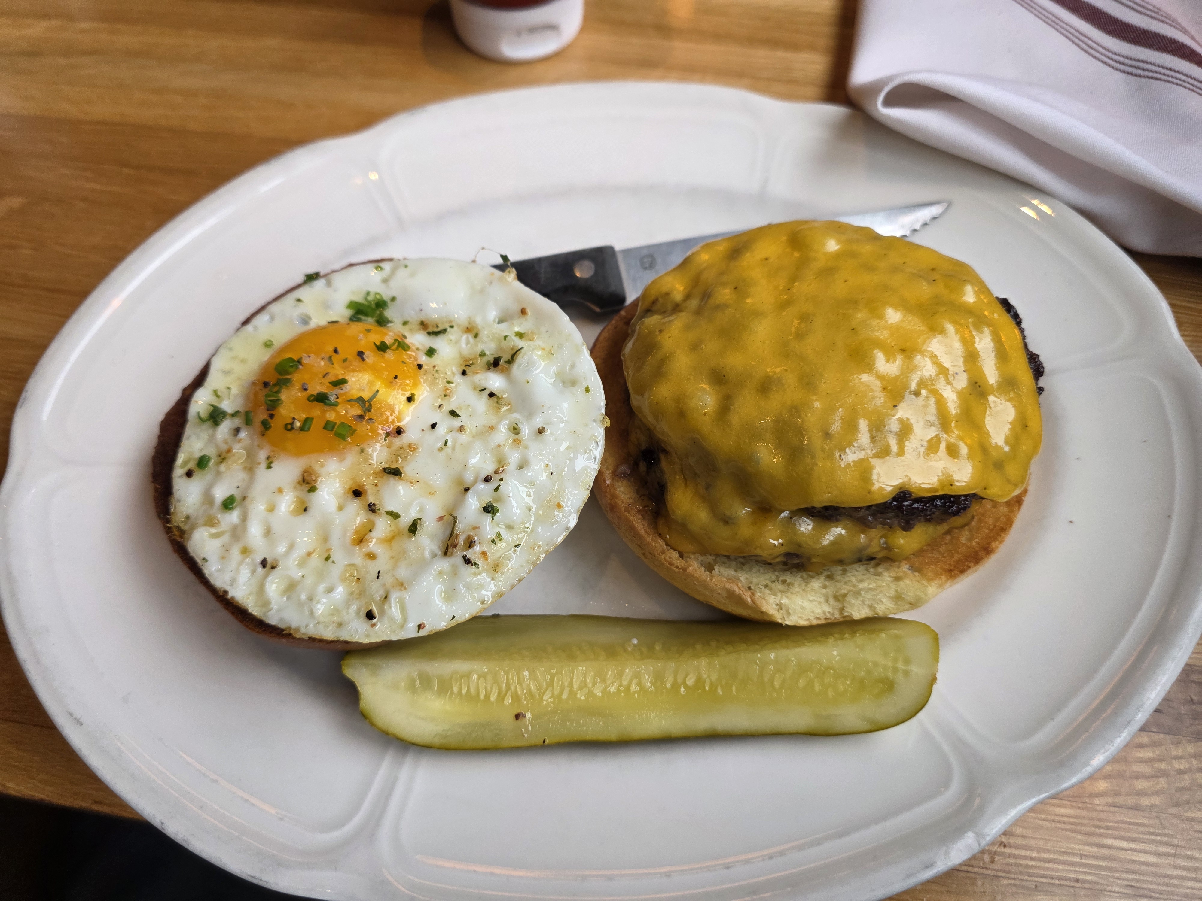 Au Cheval burger open-faced on a white plate: fried egg on one half, thick melted American cheese on the patty, cornichon alongside